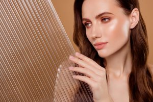 Woman with smooth, luminous skin touching a textured glass panel, illustrating the hydrated “glass skin” look.